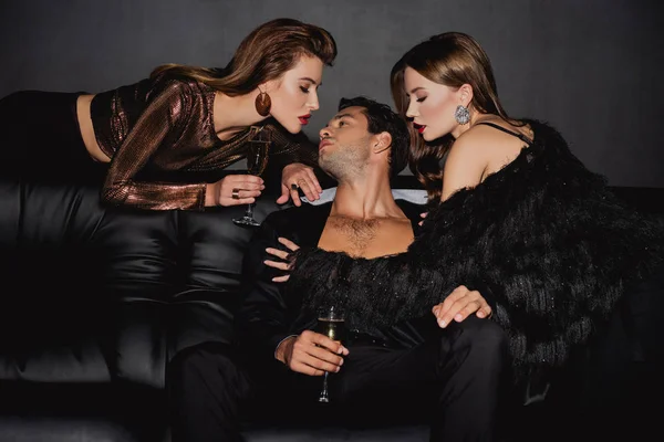Mujeres atractivas besándose hombre guapo con copa de champán aislado en negro - foto de stock