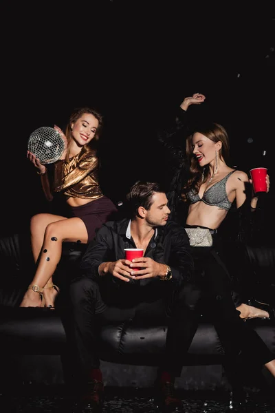 Amigos atraentes e sorridentes segurando bola de discoteca e copos de plástico isolados em preto — Fotografia de Stock