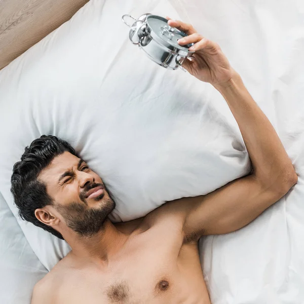 Top view of sexy and handsome bi-racial man waking up and holding alarm clock in morning — Stock Photo