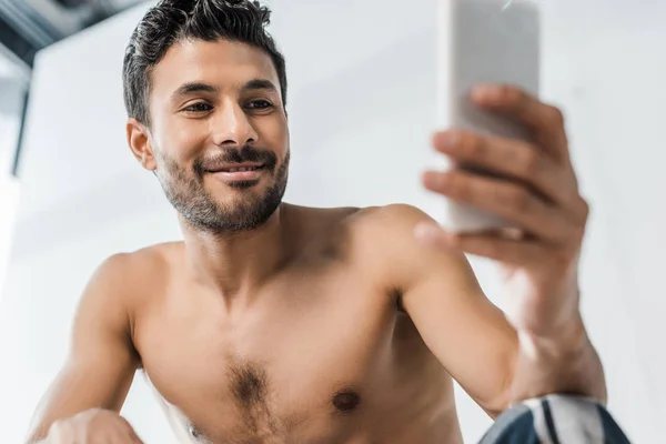 Selective focus of handsome and sexy bi-racial man smiling and using smartphone in morning — Stock Photo