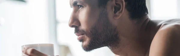 Plano panorámico de hombre bi-racial guapo sosteniendo taza de café en la mañana - foto de stock