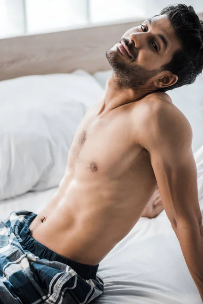 Handsome and smiling bi-racial man sitting on bed and looking at camera in morning — Stock Photo