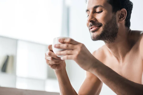 Guapo y sonriente hombre bi-racial sosteniendo taza de café en la mañana - foto de stock