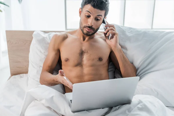 Красивый и шокированный двурасовый мужчина, указывающий рукой на ноутбук и разговаривающий по утрам на смартфоне — стоковое фото