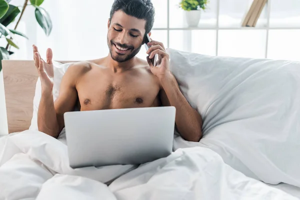 Handsome and smiling bi-racial man with laptop talking on smartphone in morning — Stock Photo