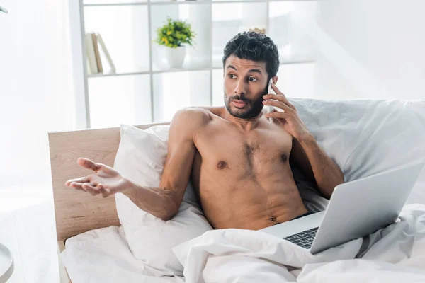 Homme bi-racial beau et choqué avec ordinateur portable parlant sur smartphone le matin — Photo de stock