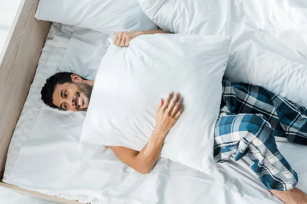 Top view of handsome and smiling bi-racial man lying on bed with pillow in morning — Stock Photo