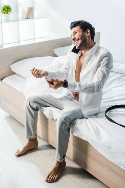 Smiling and handsome bi-racial man talking on smartphone and sitting on bed in morning — Stock Photo