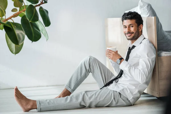 Guapo y sonriente empresario bi-racial sosteniendo taza de café en la mañana - foto de stock