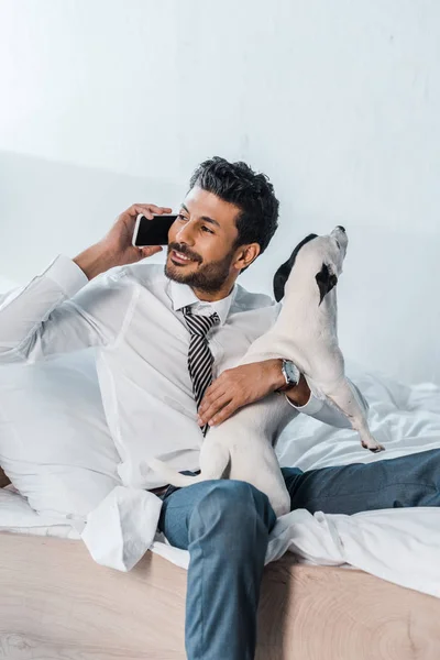 Hombre de negocios bi-racial sosteniendo Jack Russell Terrier y hablando en el teléfono inteligente en la mañana — Stock Photo