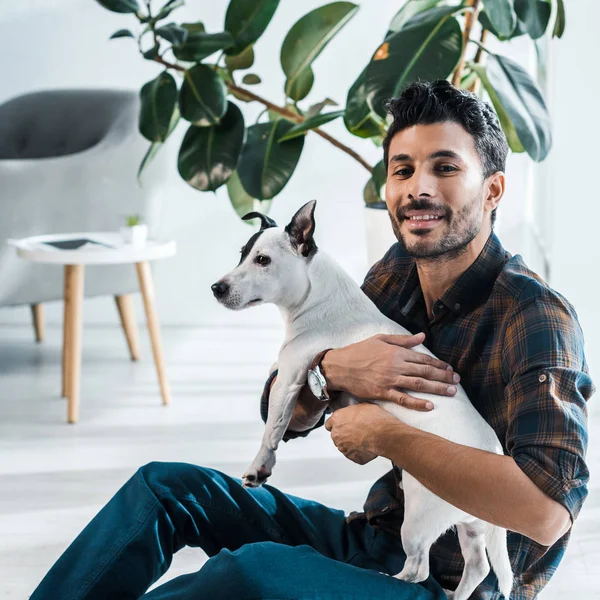 Sonriente y guapo bi-racial hombre sosteniendo Jack Russell Terrier - foto de stock