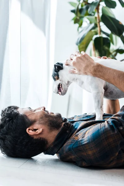 Handsome and smiling bi-racial man stroking jack russell terrier and lying on floor — Stock Photo