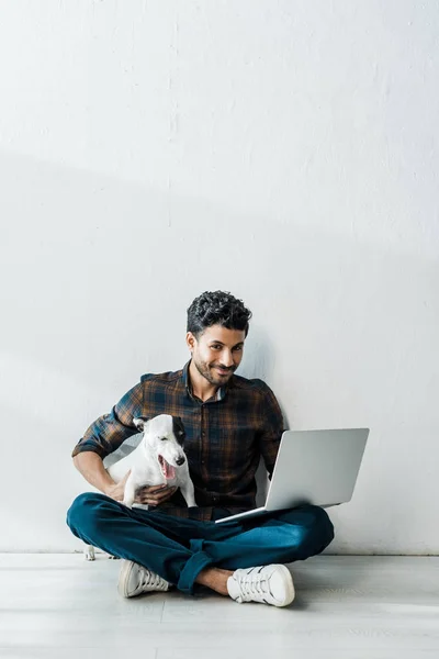 Bel homme bi-racial souriant avec ordinateur portable tenant jack russell terrier — Photo de stock