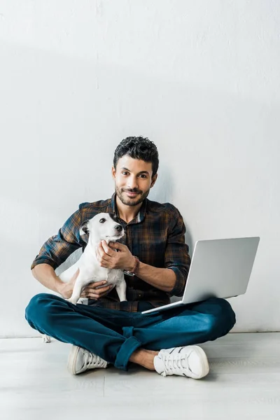 Bel homme bi-racial souriant avec ordinateur portable tenant jack russell terrier — Photo de stock