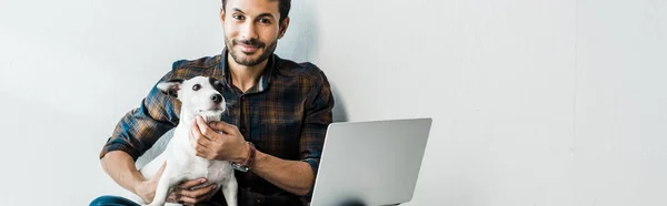 Panoramic shot of handsome and smiling bi-racial man with laptop holding jack russell terrier — Stock Photo