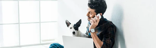 Panoramic shot of handsome bi-racial man talking on smartphone and holding jack russell terrier — Stock Photo