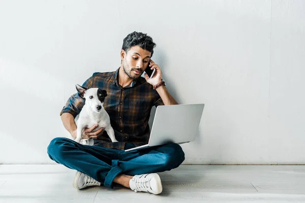 Guapo bi-racial hombre hablando en smartphone y sosteniendo jack russell terrier - foto de stock