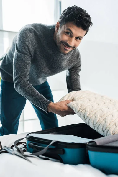 Красивый и улыбающийся двурасовый мужчина в свитере упаковывает дорожную сумку в квартиру — стоковое фото