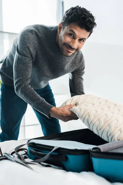 Homme bi-racial beau et souriant en pull sac de voyage d'emballage dans l'appartement — Photo de stock