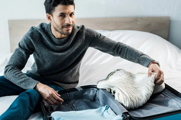 Красивый и улыбающийся двурасовый мужчина в свитере упаковывает дорожную сумку в квартиру — стоковое фото