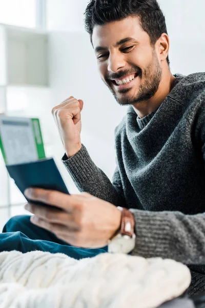 Messa a fuoco selettiva di sorridente uomo bi-razziale in maglione in possesso di passaporto con biglietto aereo e mostrando sì gesto — Foto stock