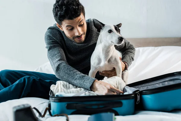 Handsome and smiling bi-racial man packing luggage and holding jack russell terrier — Stock Photo