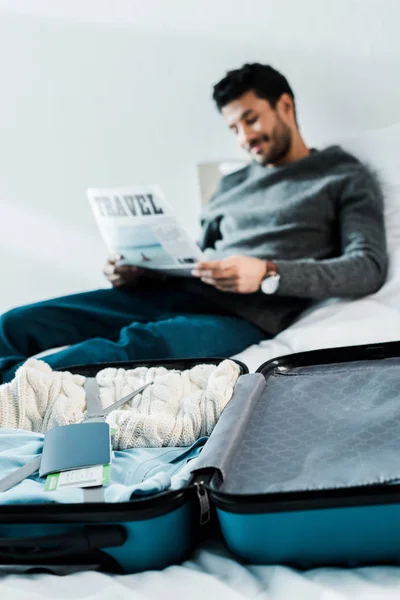 Enfoque selectivo de la bolsa de viaje y sonriente bi-racial hombre leyendo periódico con letras de viaje - foto de stock