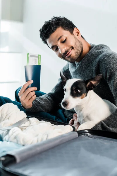 Smiling bi-racial man looking at jack russell terrier and holding passport with air ticket — Stock Photo