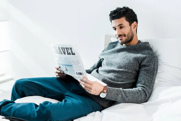 Handsome and smiling bi-racial man reading newspaper with lettering travel — Stock Photo