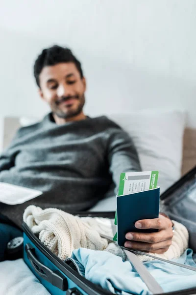 Selective focus of handsome and smiling bi-racial man holding passport with air ticket — Stock Photo