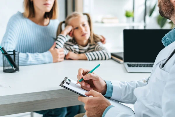 Cropped view of doctor writing prescription on clipboard near mother and daughter — Stock Photo