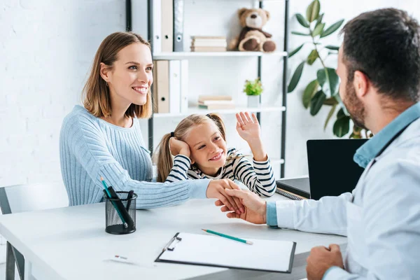 Lächelnde Frau beim Händeschütteln mit Arzt in der Nähe Tochter winkt — Stockfoto