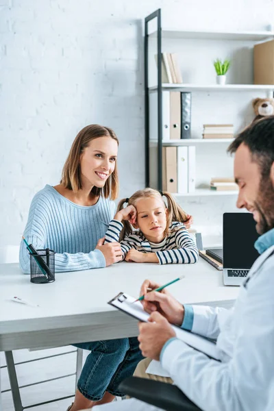 Médico positivo escribir prescripción cerca sonriente madre e hija - foto de stock