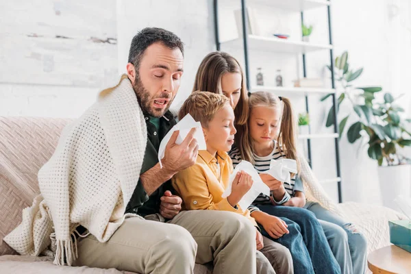 Famiglia malata, avvolta in una coperta, seduta sul divano e starnutisce nei tovaglioli — Foto stock