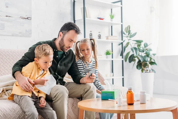 Upset man holding pills while sitting on sofa near diseased children — Stock Photo