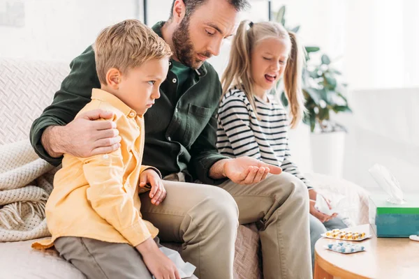 Upset man holding pills while sitting near displeased, sick children — Stock Photo