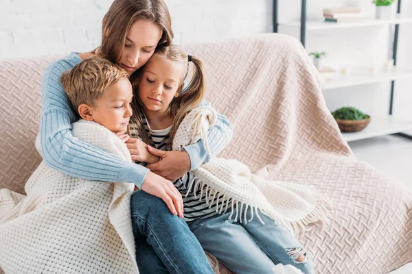 Madre cariñosa envolviendo a los niños enfermos en manta mientras está sentada en el sofá - foto de stock