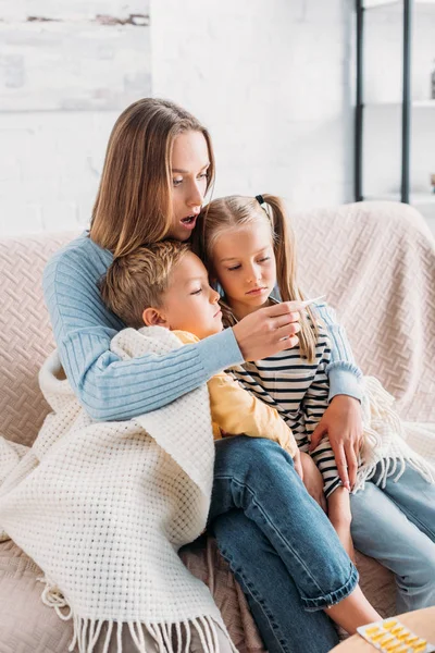 Schockierte Frau blickt auf Thermometer, während sie mit kranken Kindern auf Sofa sitzt — Stockfoto