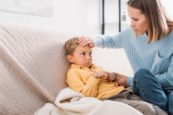 Worried woman touching forehead of sick son sitting on sofa — Stock Photo