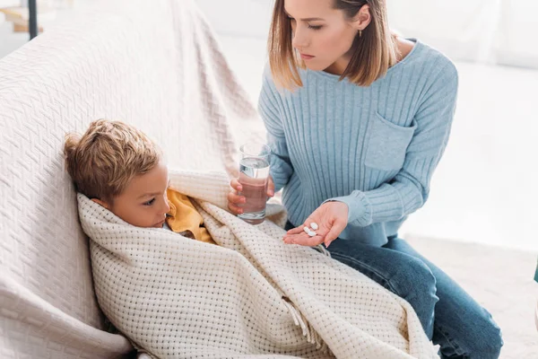 Attentive mother giving medicines to diseased son wrapped in blanket — Stock Photo