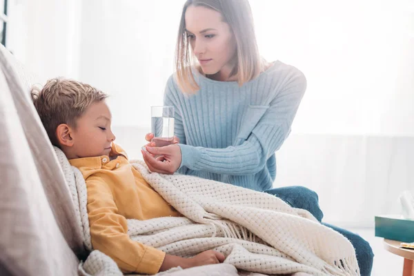 Attentive mother giving medicines to ill son wrapped in blanket — Stock Photo