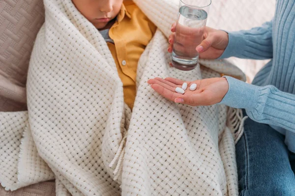 Vue recadrée de la femme tenant des pilules et un verre d'eau près du fils malade — Photo de stock