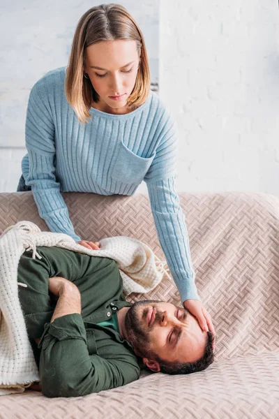 Уважна дружина торкається лоба хворого чоловіка, що лежить на дивані — стокове фото