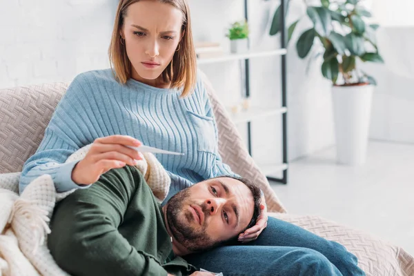 Aufmerksame Ehefrau blickt auf Thermometer neben krankem Mann, der auf dem Schoß liegt — Stockfoto