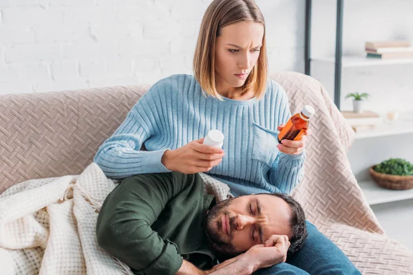 Aufmerksame Ehefrau hält Behälter mit Tabletten und Hustensaft neben krankem Mann auf dem Schoß — Stockfoto