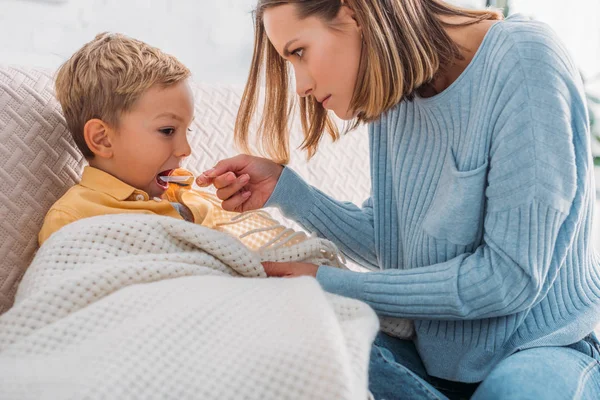 Mãe atenta dando remédio para o filho doente envolto em cobertor — Fotografia de Stock