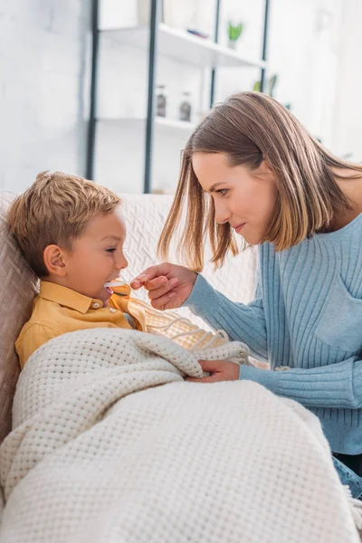 Smiling woman giving medicine to diseased son wrapped in blanket — Stock Photo