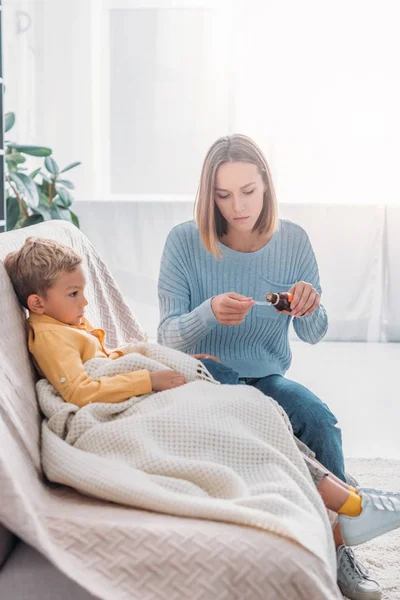 Attentive mother pouring cough syrup in spoon while sitting near sick son — Stock Photo