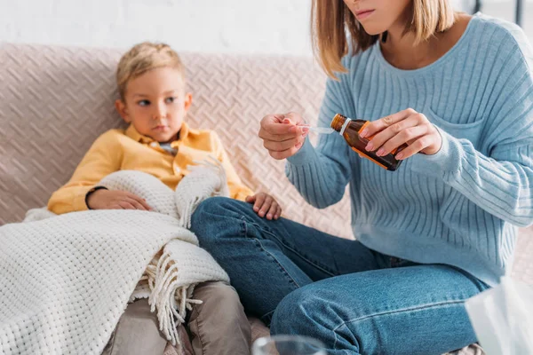 Vue recadrée de la mère versant du sirop contre la toux dans une cuillère alors qu'elle était assise près d'un fils malade — Photo de stock