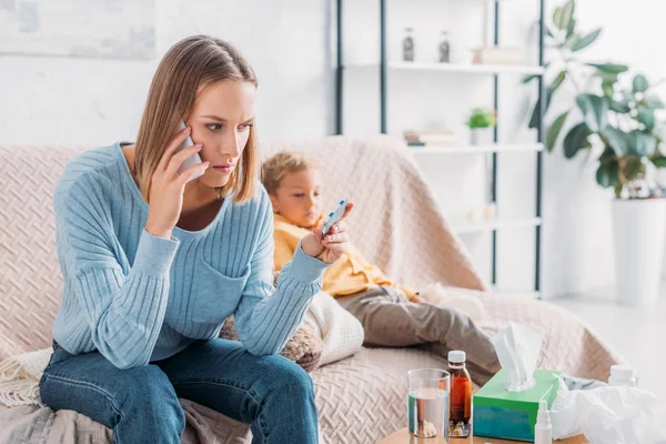 Madre preocupada sosteniendo ampolla de píldoras y hablando en el teléfono inteligente mientras está sentado cerca de hijo enfermo - foto de stock